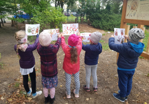 Grupa dzieci stoi w ogrodzie, ogląda swoje prace plastyczne i szuka podobnych barw w otoczeniu.